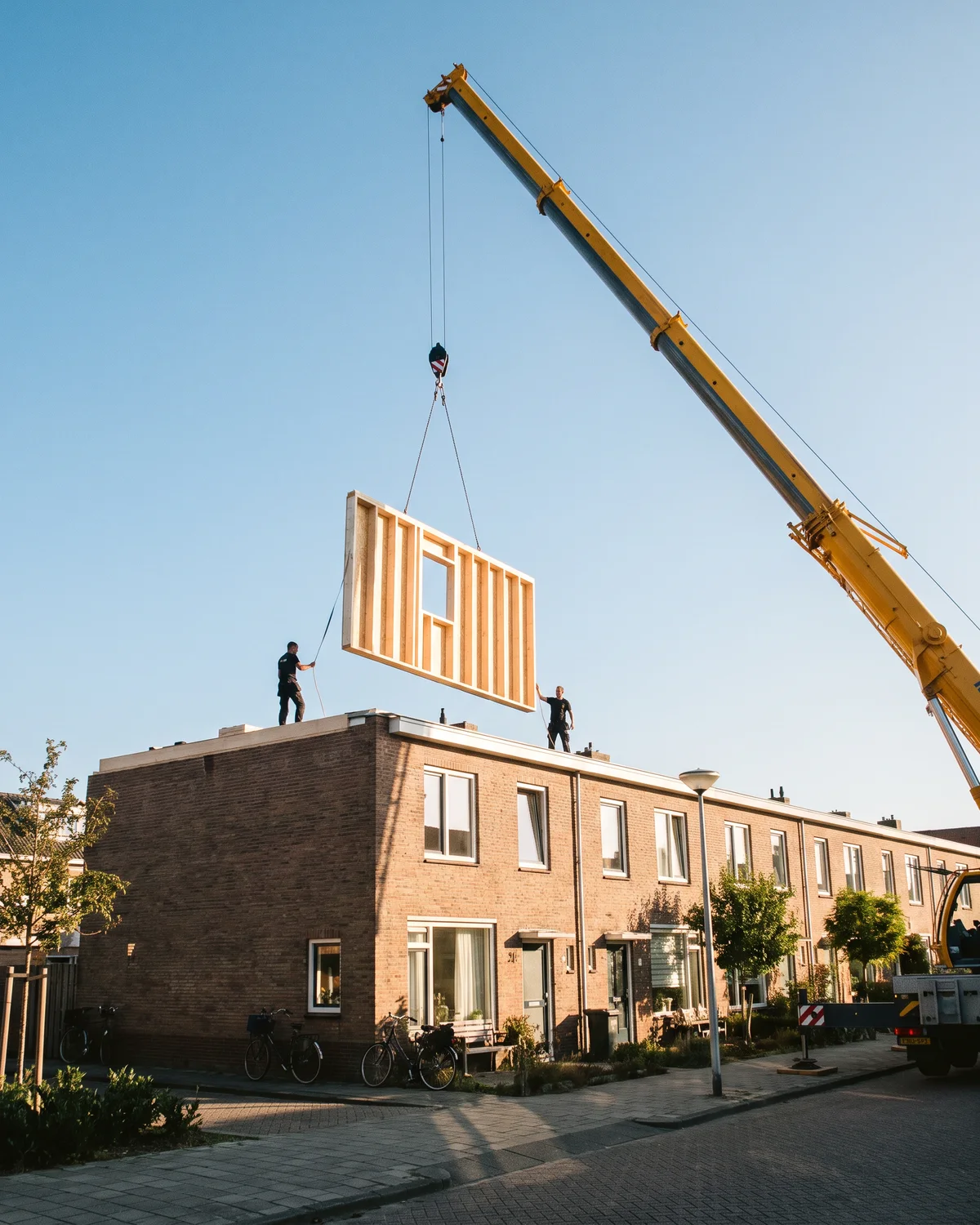 Een kraan hijst een prefab wand van de opbouw boven het platte dak; twee monteurs staan klaar