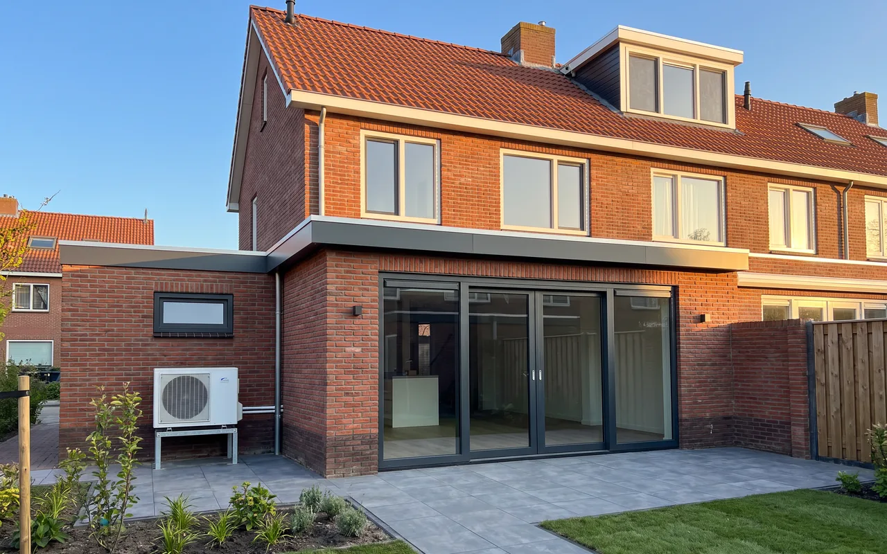 Achtergevel van een rijwoning met een nieuwe uitbouw, geïsoleerd plat dak en een warmtepomp tegen de gevel, in de middagzon